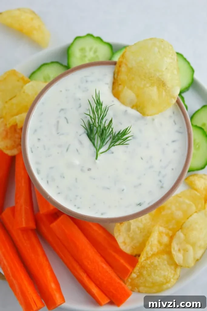 Greek yogurt dill dip displayed in white bowl on a plate, surrounded by chopped vegetables and potato chips. A potato chip is placed in the bowl with the dip.