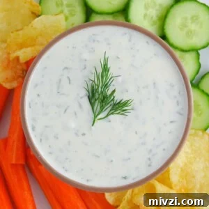 Greek yogurt dill pickle dip presented in a brown bowl on a plate with chopped veggies and potato chips. Food is on a white marble countertop.