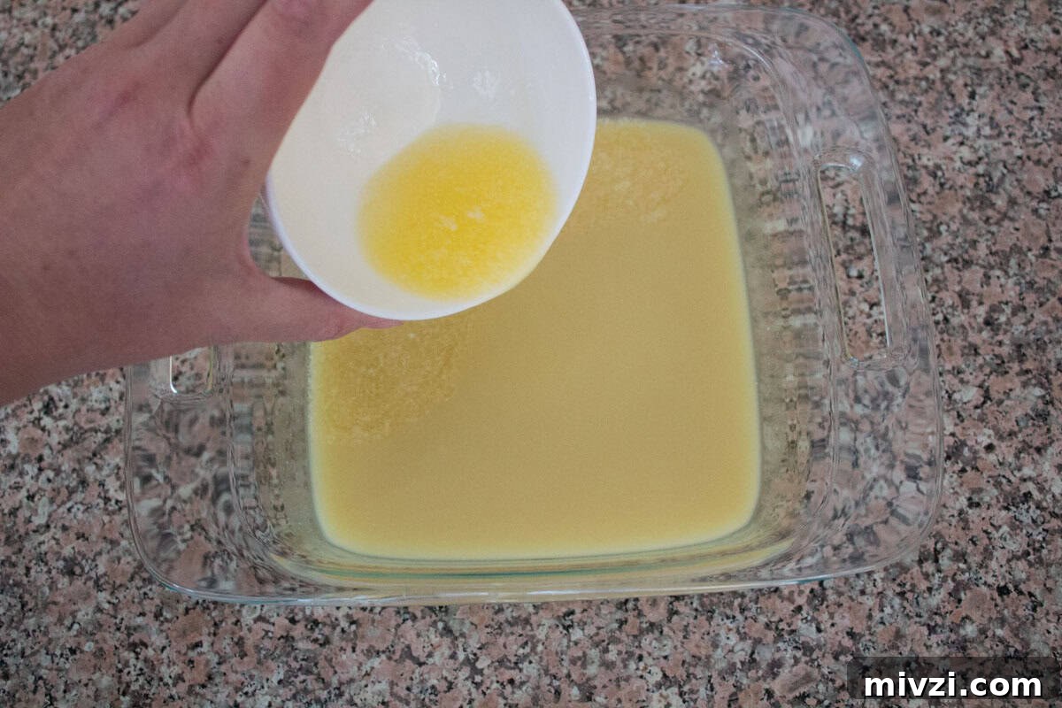 A hand pours melted butter from a white bowl into a glass baking dish filled with yellow liquid, on a speckled countertop.