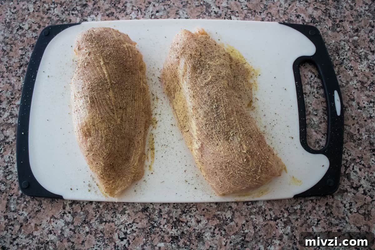 Two raw, seasoned chicken breasts on a white cutting board with black edges, placed on a speckled brown countertop.