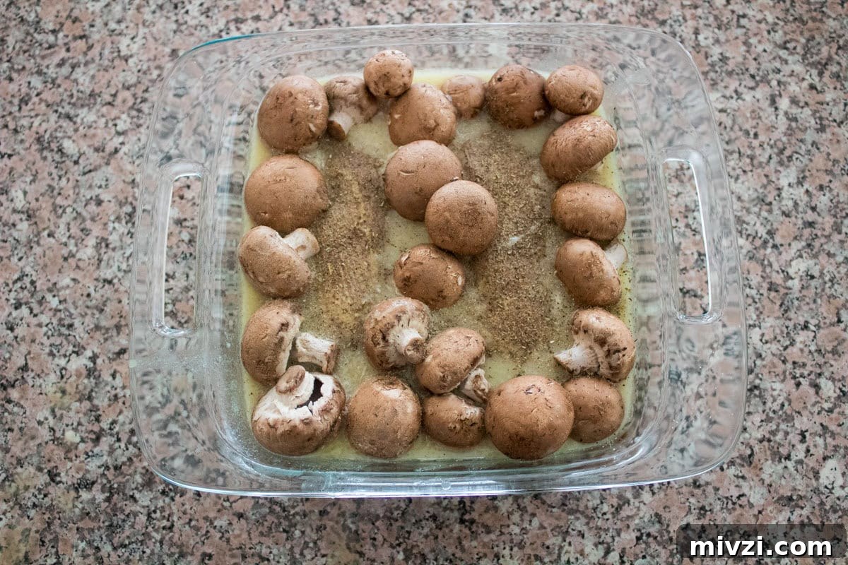 A glass baking dish filled with whole brown mushrooms arranged along the edges, sitting in a pale liquid with seasoning, on a granite countertop.