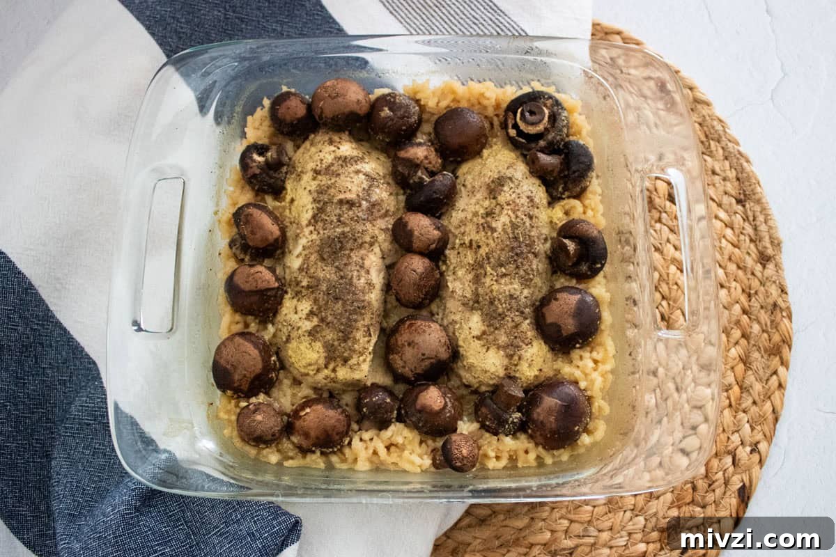 A glass baking dish with two seasoned chicken breasts, whole mushrooms, and rice, placed on a woven placemat next to a folded kitchen towel.
