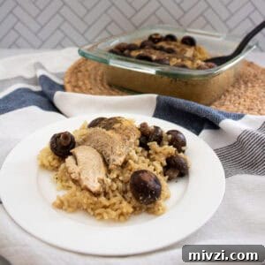 A plate with baked chicken breast, brown rice, and mushrooms sits in the foreground, while a glass baking dish with more of the same food is in the background on a woven mat. The setting includes a striped cloth.