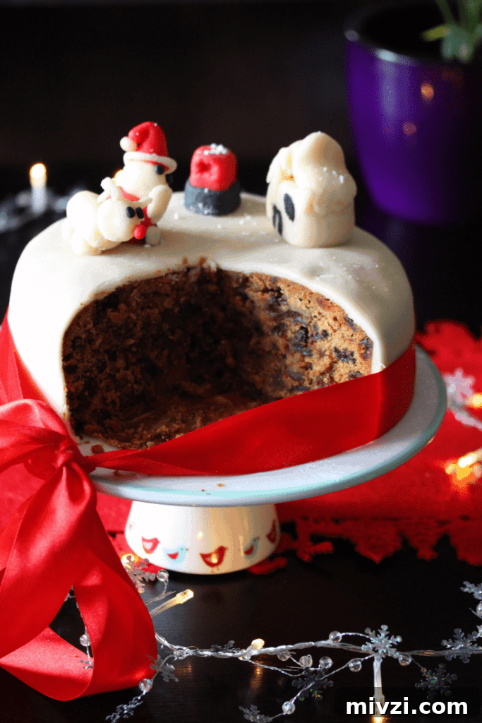 Dairy-Free Gluten-Free Christmas Cake Recipe 4 A close up of an iced Dairy Free Christmas Cake on a stand and surrounded by festive decorations. Several slices have been removed to show the dark fruity interior of the cake.