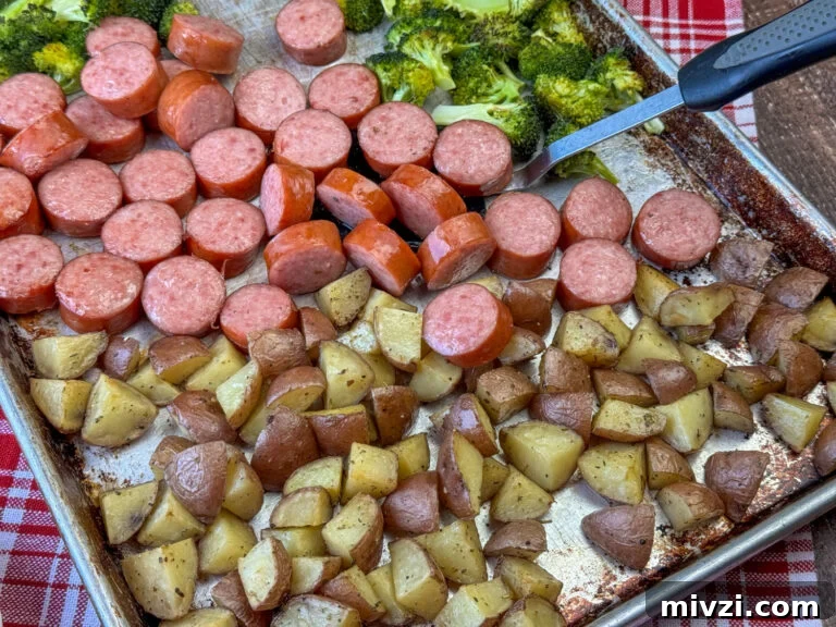 Spatula scooping browned roasted Kielbasa, potatoes and broccoli off of a baking sheet.