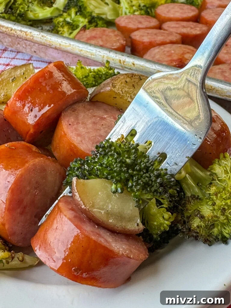 Close up of a fork full of broccoli, a potato chunk and thick piece of Kielbasa with more sausage in the background on the plate.