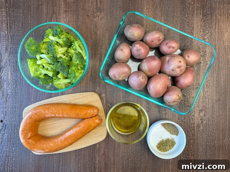 Ingredients to make kielbasa in the oven are Kielbasa sausage, baby red potatoes, olive oil, salt, pepper, Italian seasoning and broccoli on a wooden counter top.