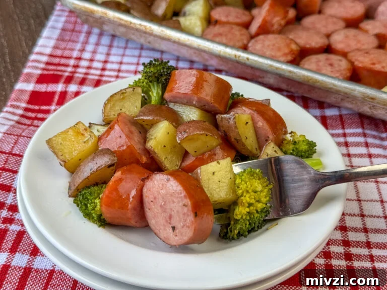 Close up of a fork full of juicy roasted Kielbasa, tender potatoes and cooked broccoli on a white plate on a red and white napkin in front of a sheet full of sausage.
