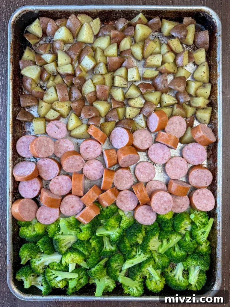 Overhead view of thick coins of Kielbasa added to a sheet pan or partially roasted potatoes and broccoli to finish cooking in the oven.