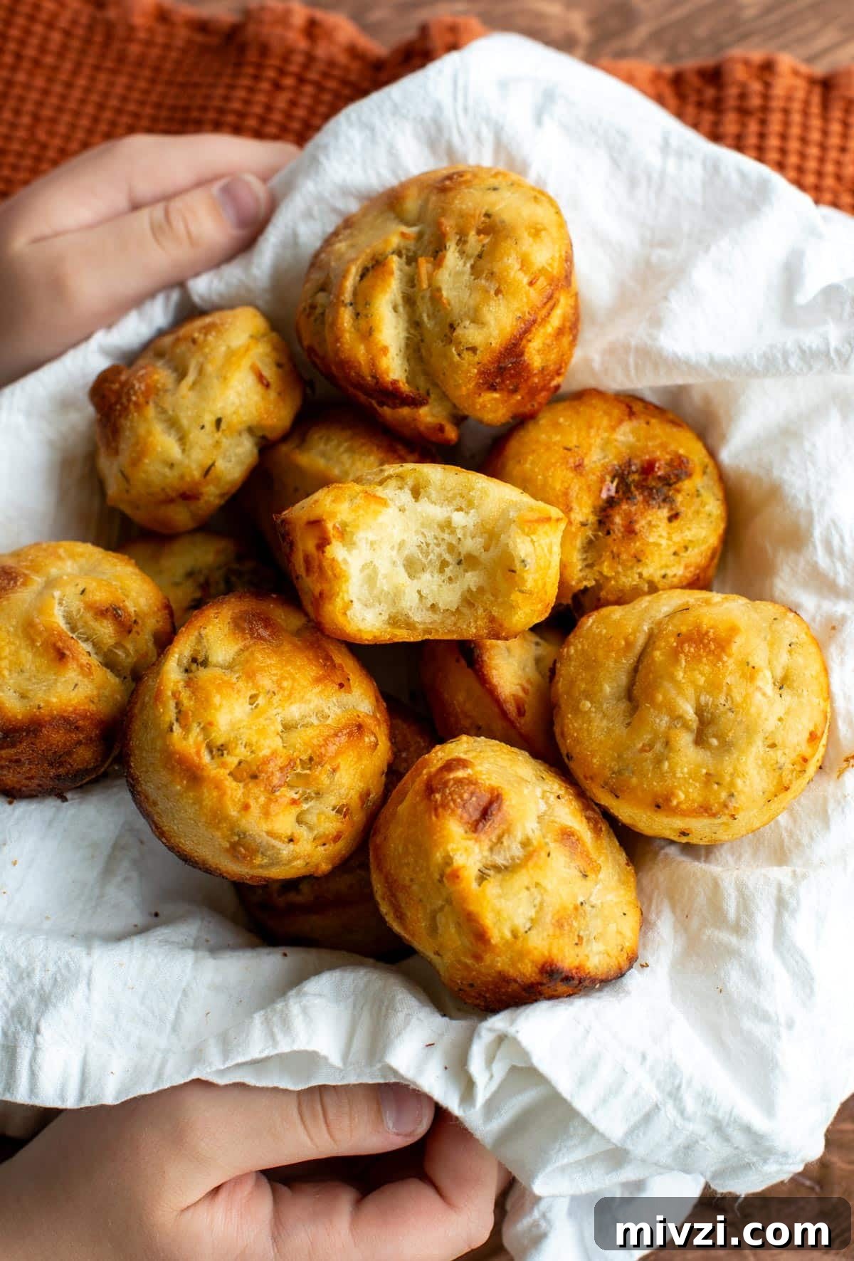 Hands holding a basket lined with a white cloth, filled with golden sourdough focaccia rolls, one torn open to reveal a soft, airy interior.