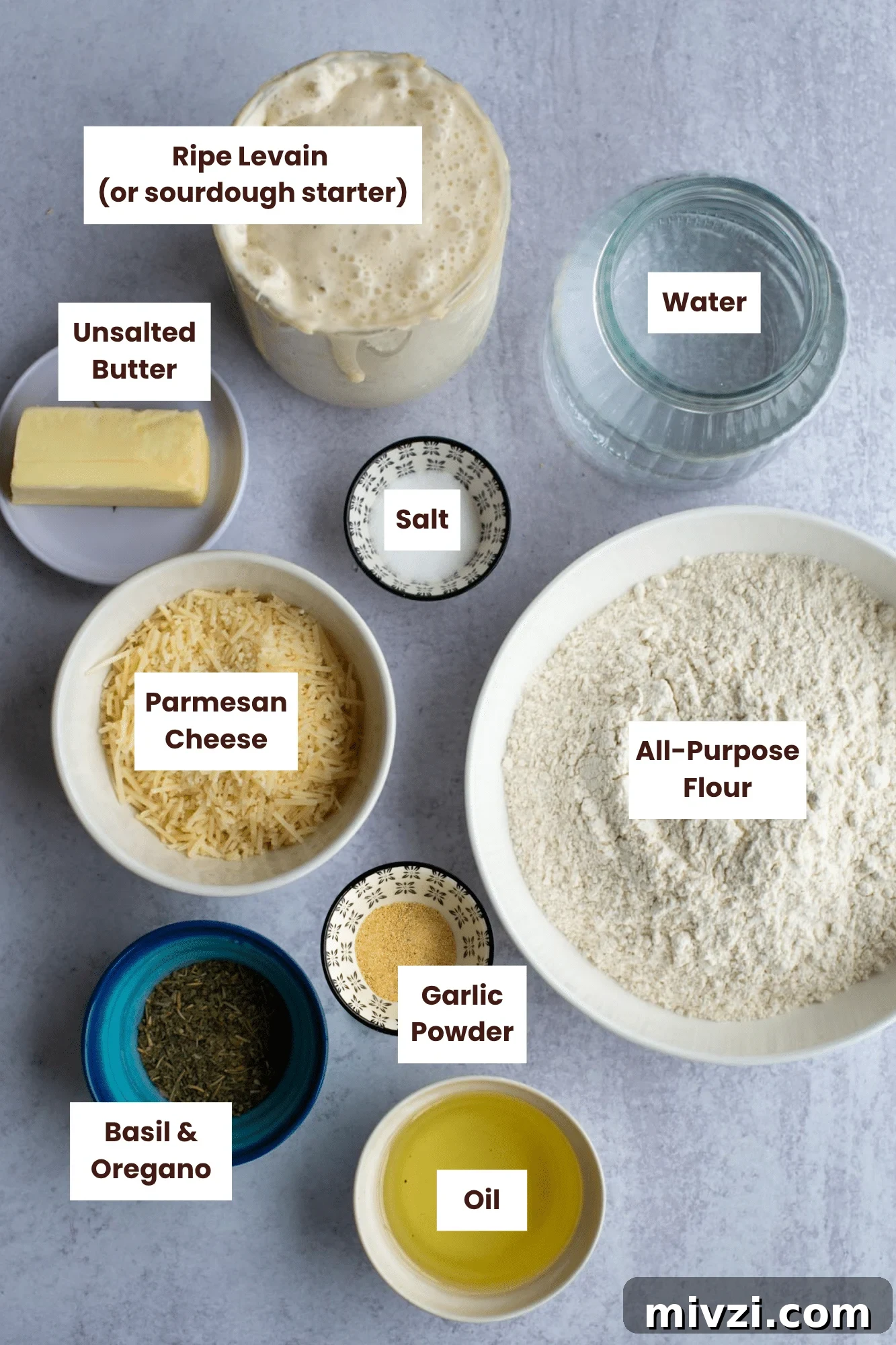 Overhead view of labeled ingredients for sourdough focaccia rolls, including ripe levain, bread flour, water, salt, butter, olive oil, Parmesan cheese, garlic, and dried oregano and basil arranged neatly in bowls.