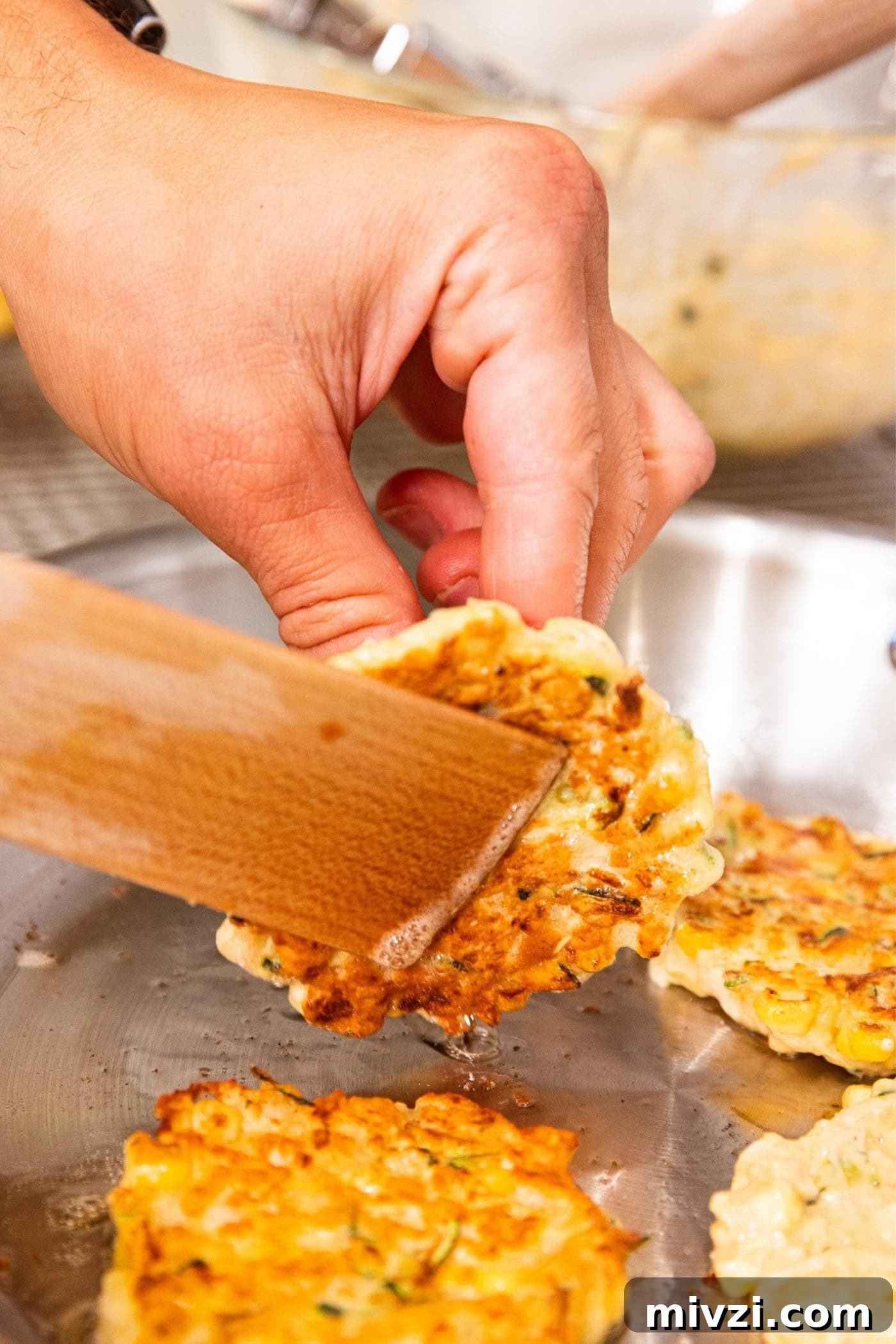 Hand using wooden spatula to flip corn fritter in metal skillet on stove.