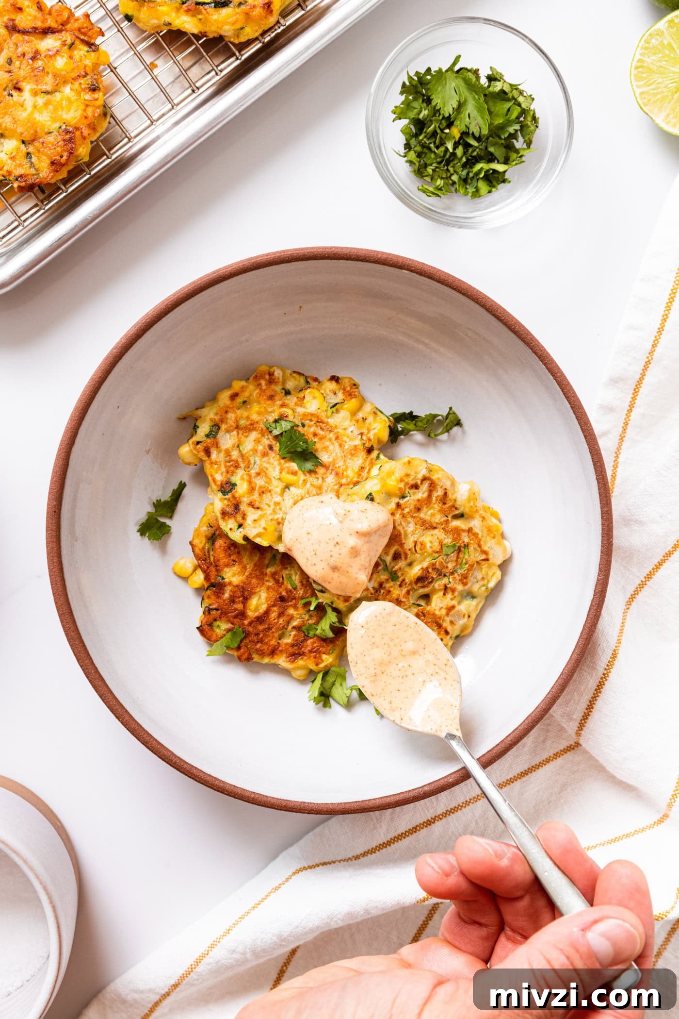 White bowl with three corn fritters topped with cilantro and spoonful of chipotle mayonnaise sauce all on white countertop.