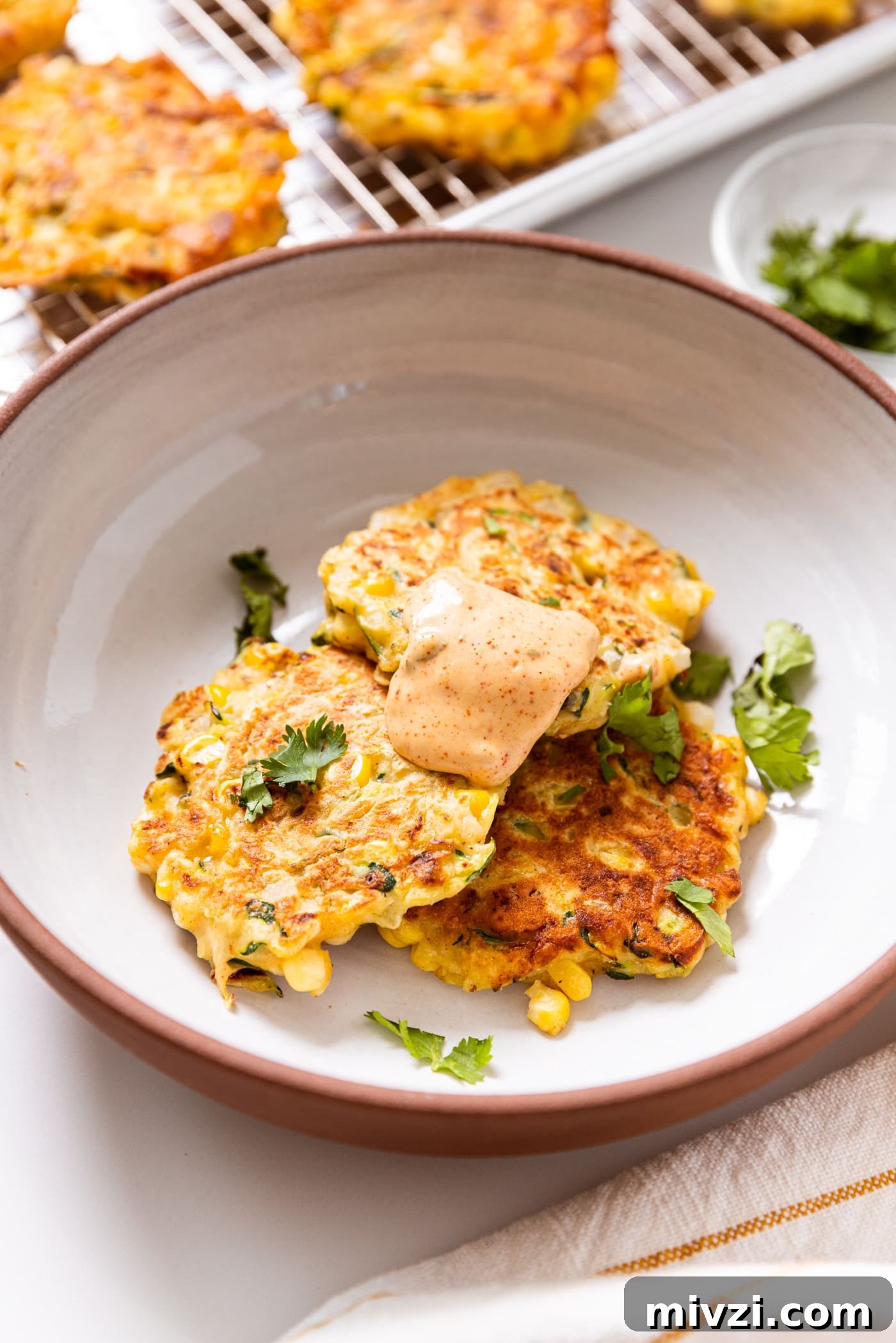 White bowl filled with three fried zucchini and corn fritters topped with chipotle mayonnaise sauce with cilantro sprinkled on top with more fritters cooling in background.