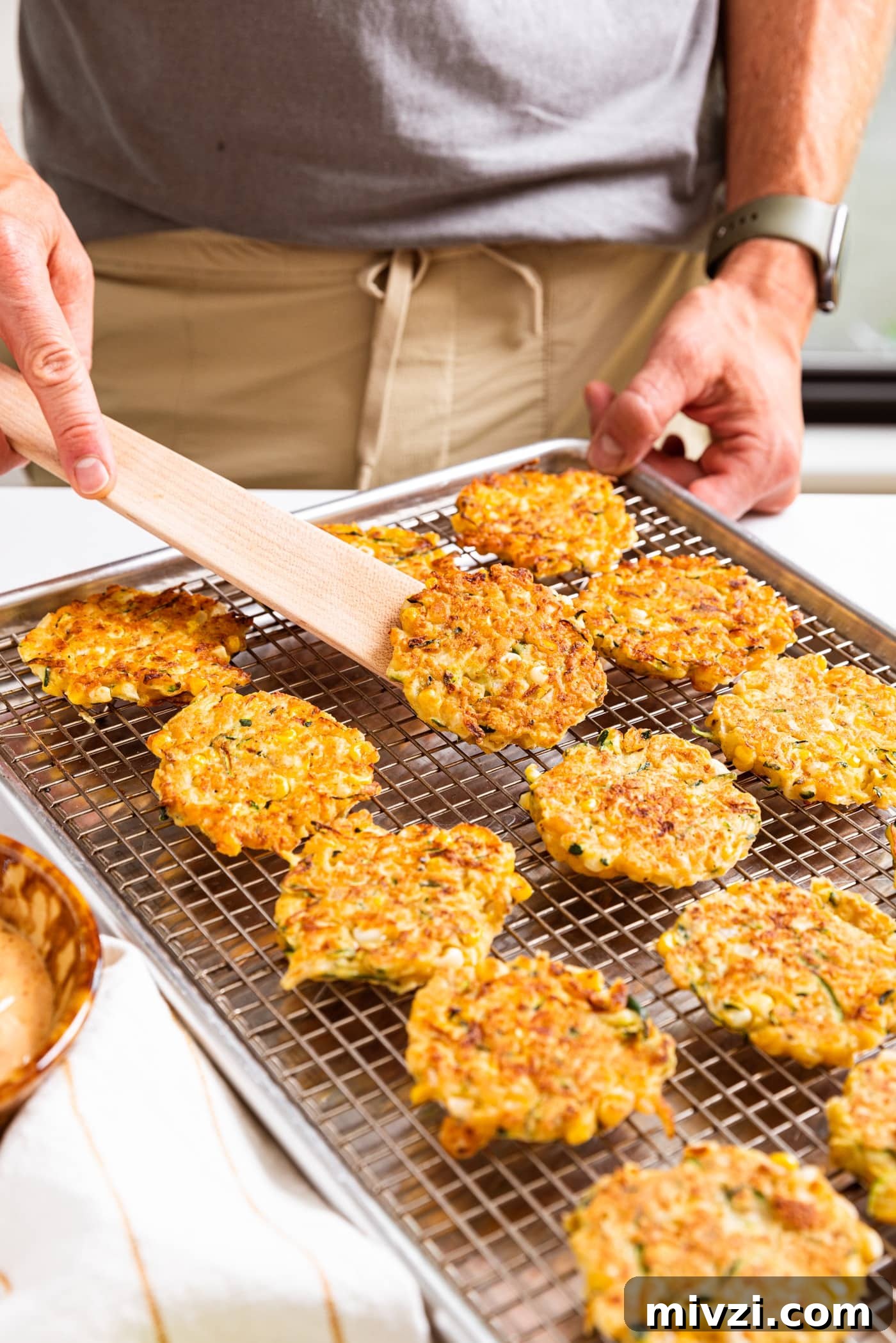 Hand holding wooden spatula scooping up one fried zucchini and corn fritter off of metal cooling rack filled with fritters all on white countertop.