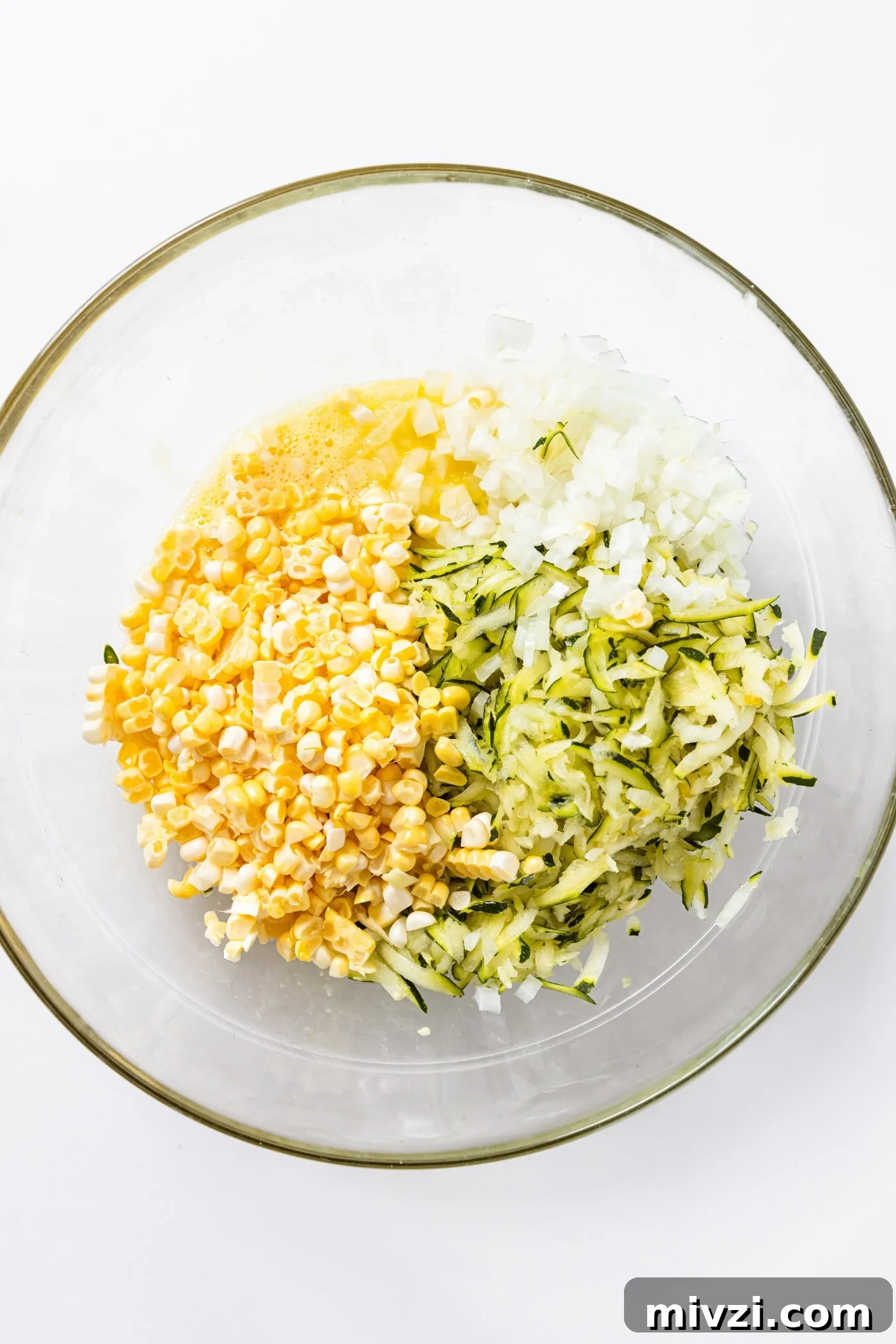 Glass bowl sitting on white countertop filled with onion, corn, and zucchini.