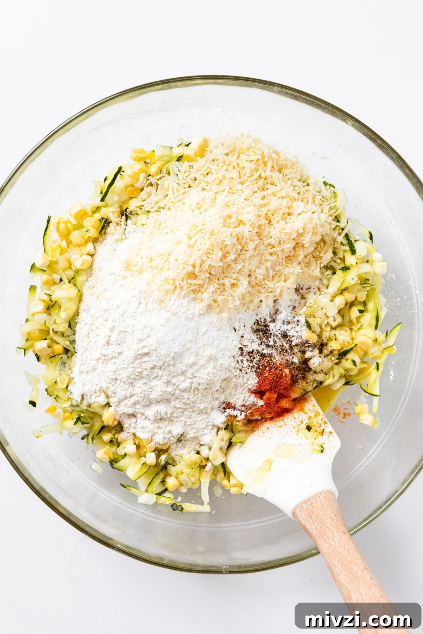 Corn and zucchini mixture sitting in glass bowl topped with flour, Parmesan cheese, and spices.