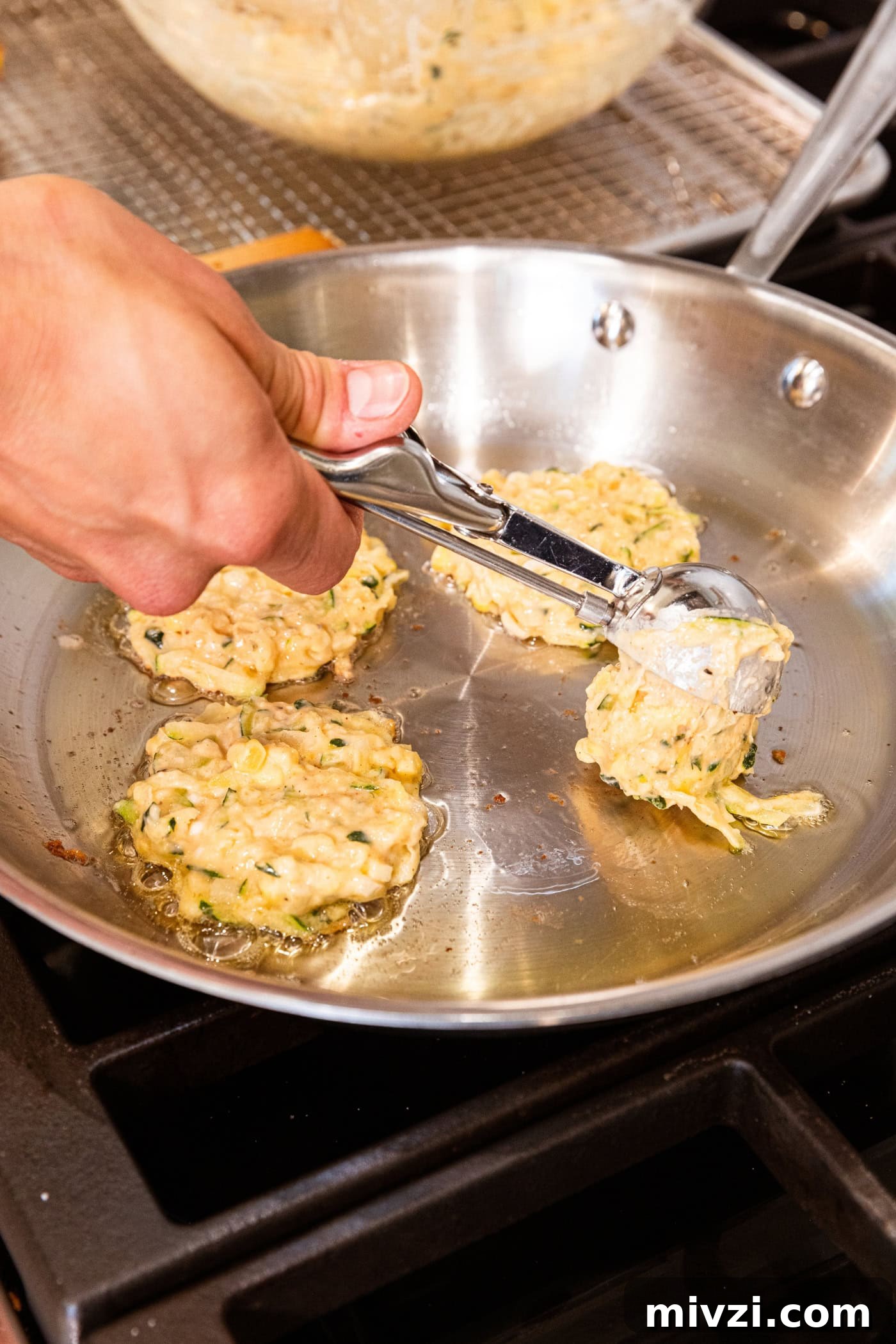Hand dropping fritter batter from ice cream scoop onto hot metal skillet on stove.