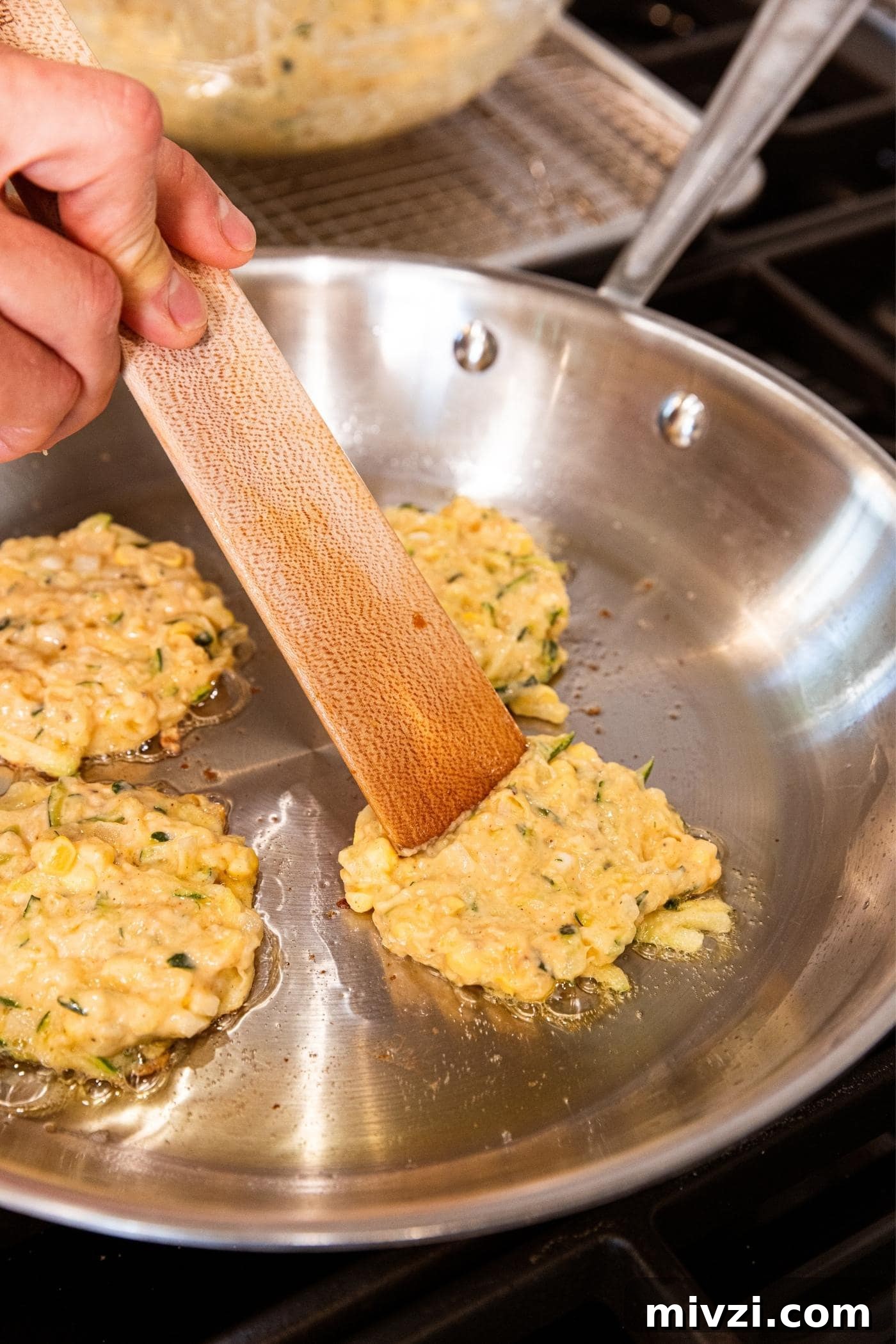 Hand pressing out fritter batter with wooden spatula on metal skillet on stove.