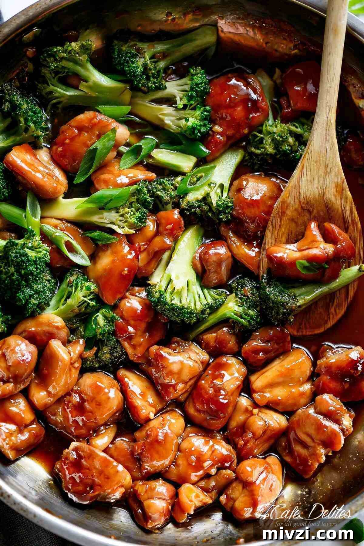 Close-up image of Teriyaki Chicken with broccoli and chives in a pan, being stirred by a wooden spoon soaked in teriyaki sauce.