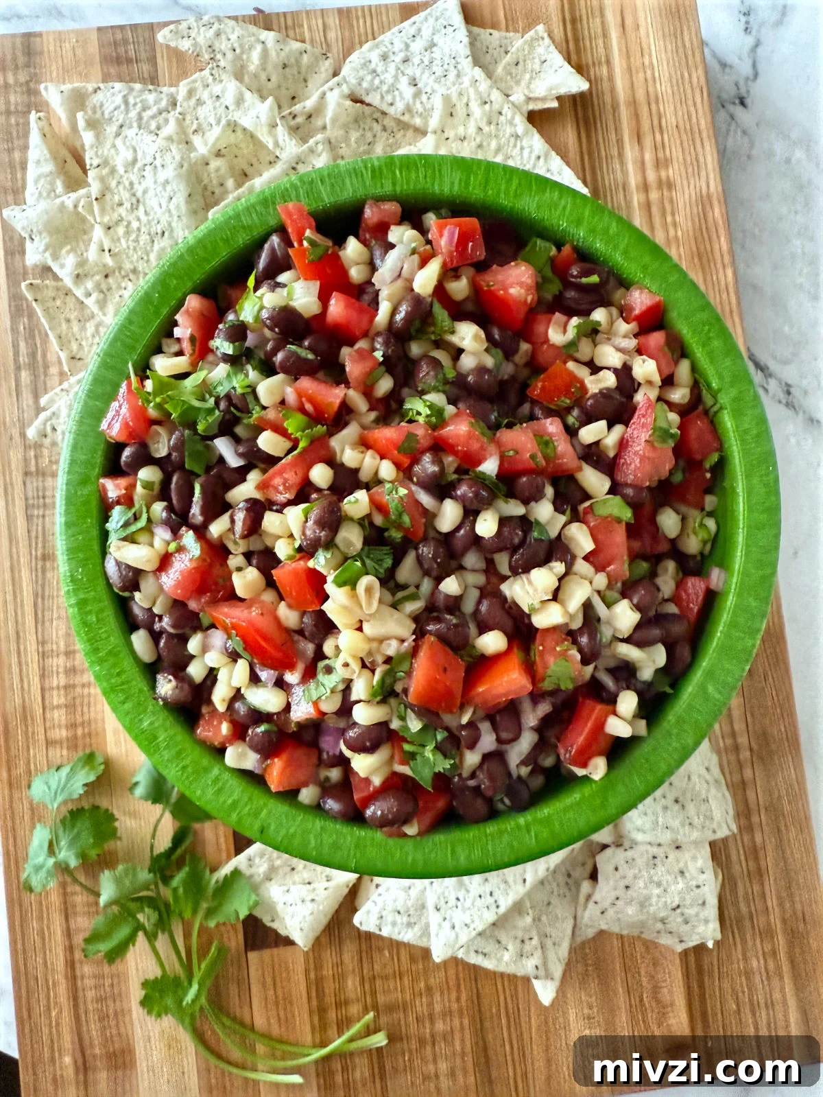 Texas Caviar: Zesty Black Bean and Corn Salsa Dip 2 Overhead view of a green bowl filled with black bean, corn, tomato, and cilantro dip, surrounded by tortilla chips on a wooden board.