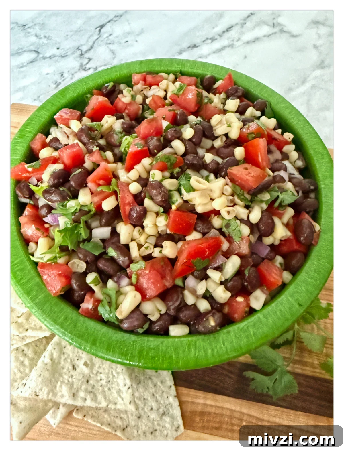 Texas Caviar: Zesty Black Bean and Corn Salsa Dip 5 Black bean and corn dip with diced tomatoes, red onion, and cilantro in a green bowl, served with tortilla chips on a wooden board with a marble background.