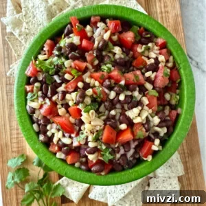 Overhead view of a green bowl filled with black bean, corn, tomato, and cilantro dip, surrounded by tortilla chips on a wooden board.