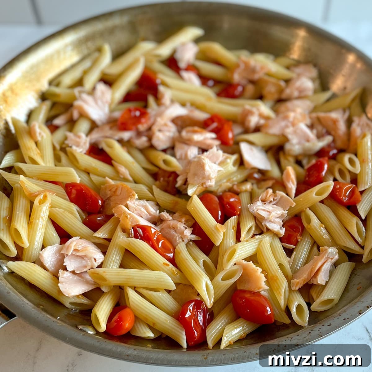 Penne pasta mixed with flaked salmon and burst cherry tomatoes in a stainless steel pan over a white marble countertop.