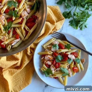 A white plate of penne pasta with burst cherry tomatoes and flaked salmon topped with parsley sits on a white marble countertop with a yellow linen, a pan of salmon penne pasta, a bunch of parsley, and a copper-colored fork.