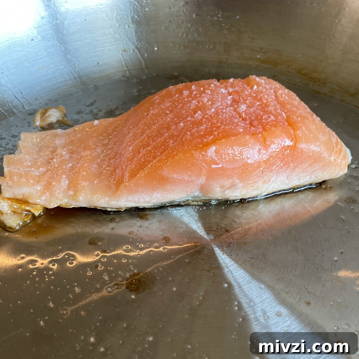 A fillet of salmon is cooking in a stainless steel pan with olive oil and salt.