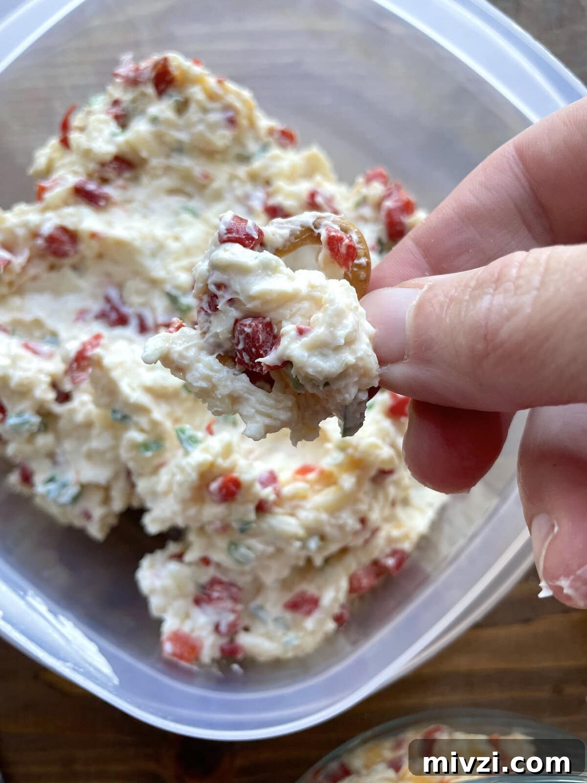 Spicy Jalapeño Pimento Cheese Dip Recipe 6 A hand holding scoop of pimento cheese with jalapeños on a pretzel over a Tupperware.