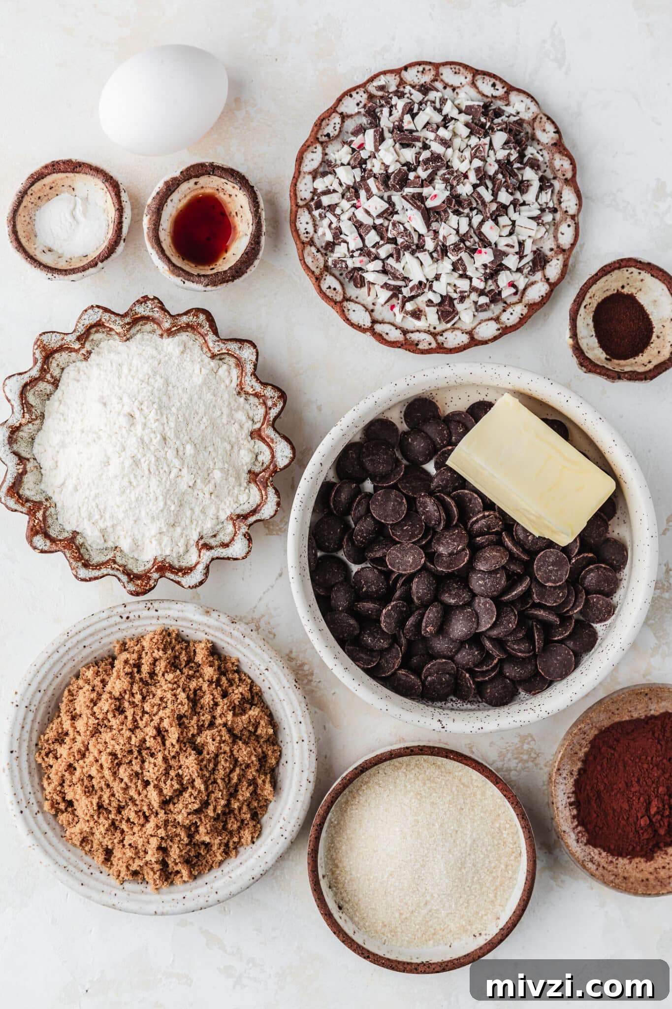 Chewy Mini Chocolate Peppermint Bark Cookies for Holiday Baking 3 White and brown bowls of chocolate chips, butter, flour, brown sugar, white sugar, cocoa powder, vanilla, baking soda, espresso powder, and peppermint candy next to an egg on a tan counter.