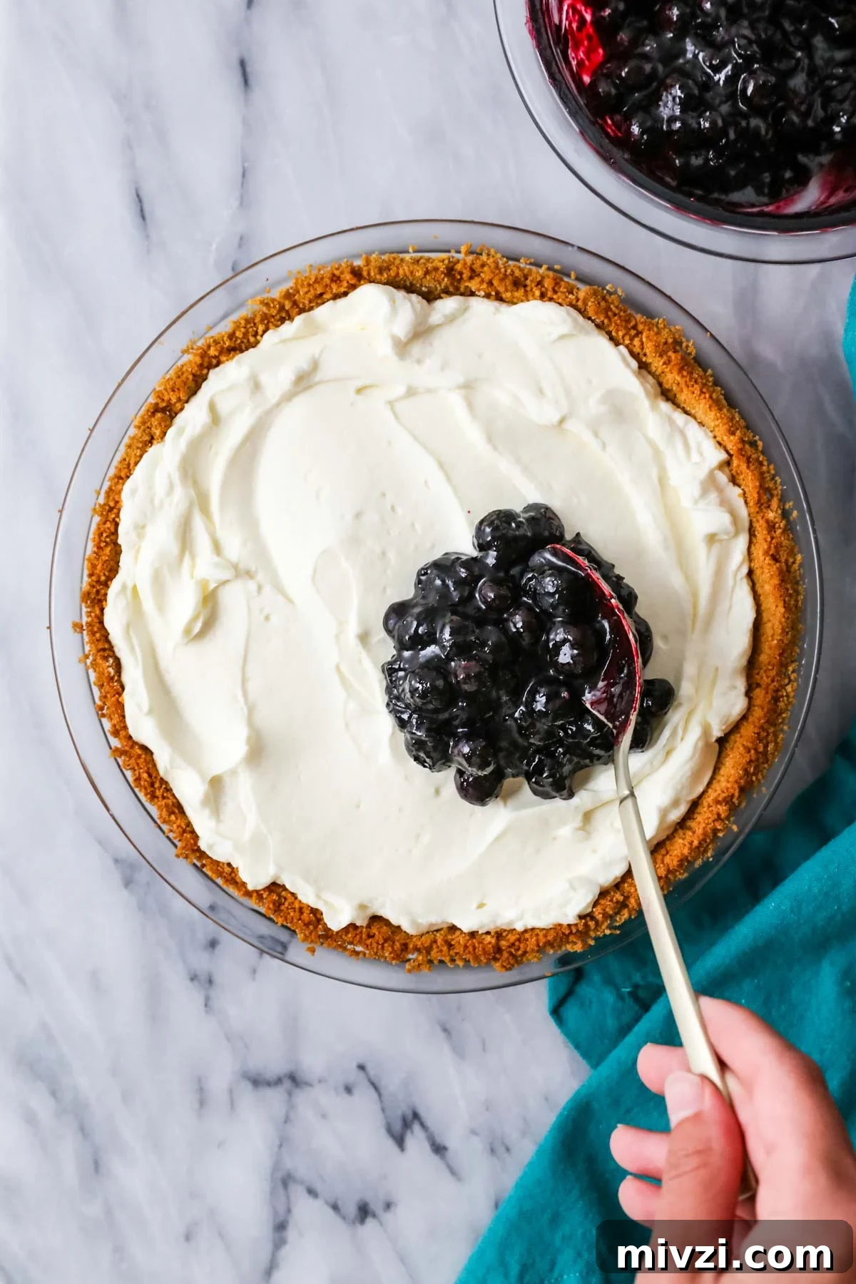 Blueberry topping being spooned over a cream cheese pie.