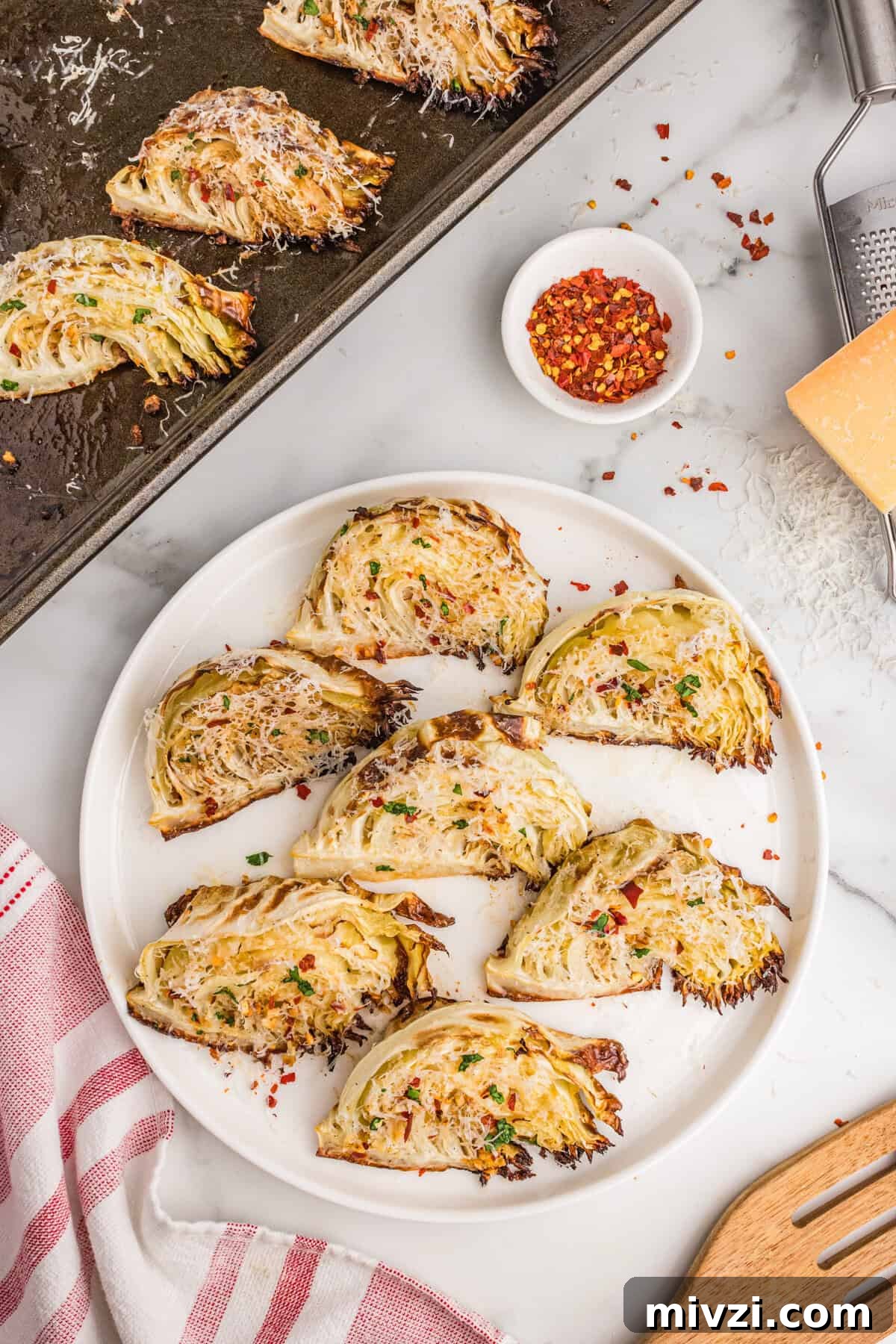 Crispy Roasted Cabbage Wedges with Garlic and Parmesan 9 Roasted garlic parmesan cabbage wedges arranged on a white serving plate and baking sheet, topped with shredded parmesan, parsley, and red pepper flakes.