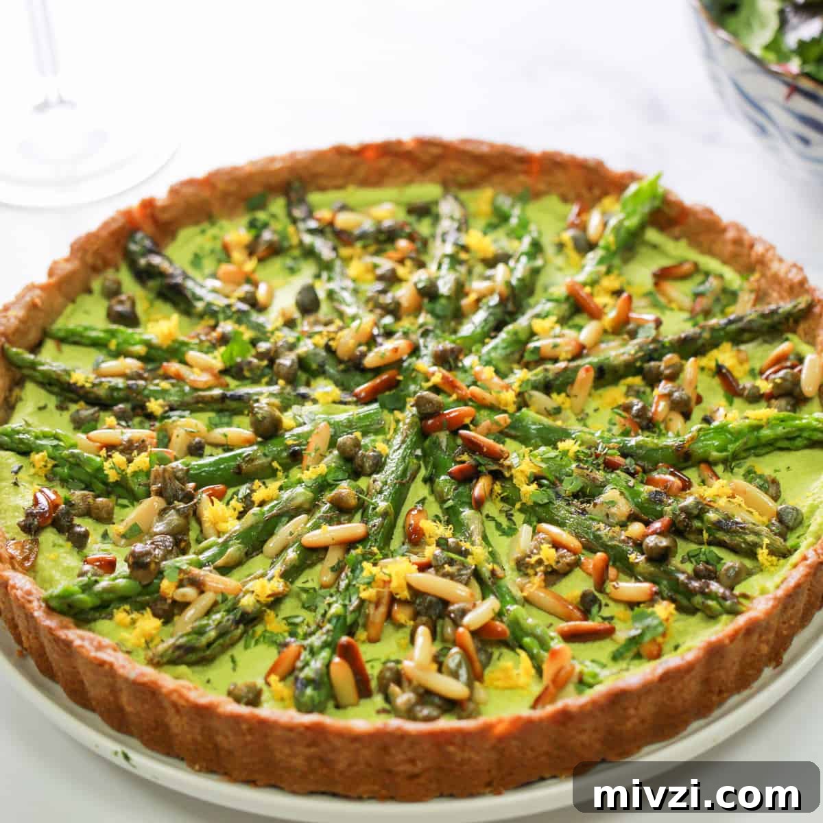Photo of a whole Asparagus and Pesto Macadamia Tart on a white marble surface with a bowl of salad in the background.