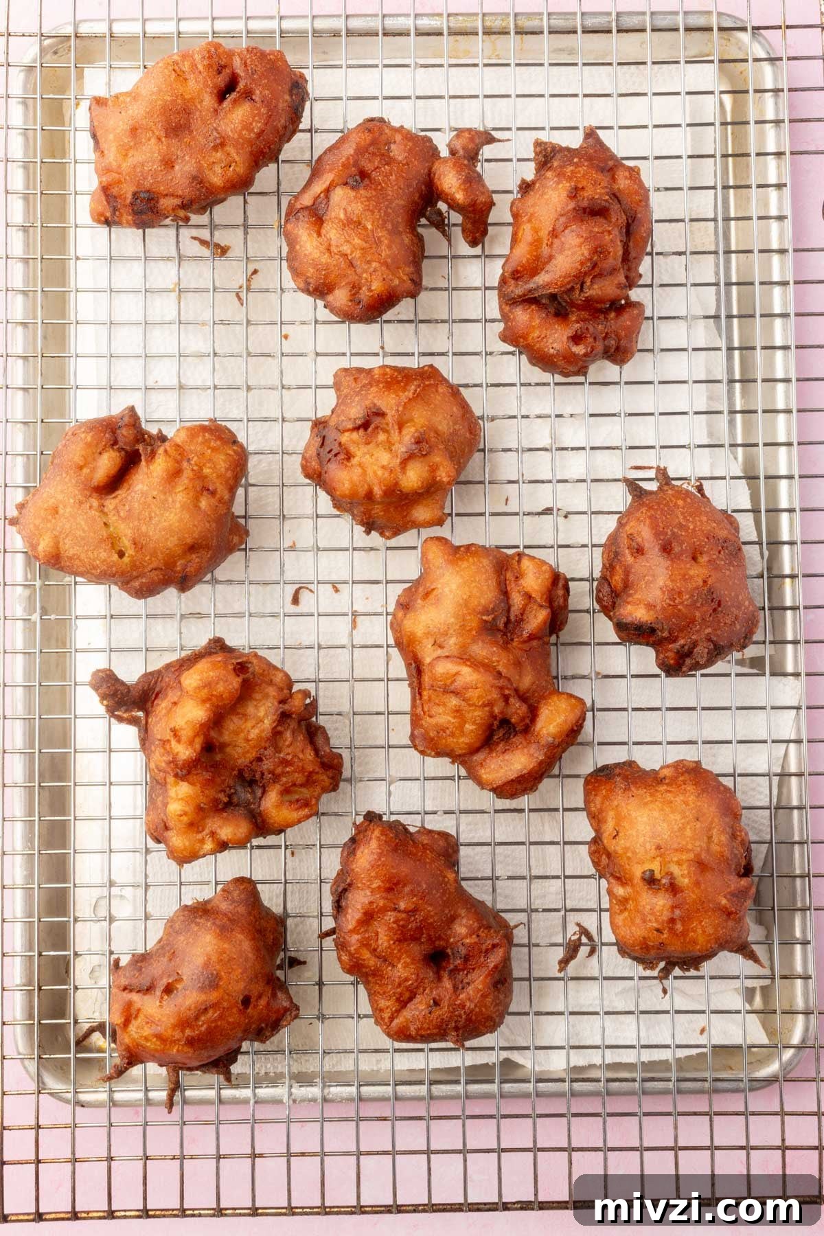 Crispy Gluten-Free Apple Fritters Recipe 12 Fried gluten-free apple fritters on a cooling rack lined with paper towels.