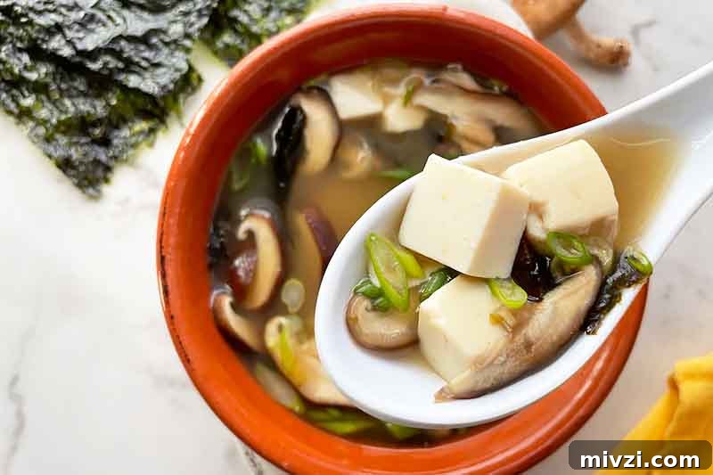 Horizontal image of spoon holding cubes of tofu with scallions and mushrooms over a bowl of broth.