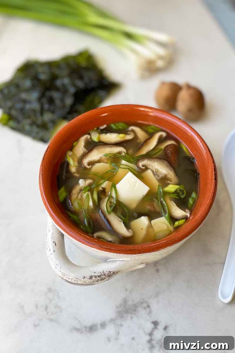 Vertical image of a red bowl with white handles filled with a broth with mushrooms and tofu cubes next to seaweed and scallions.