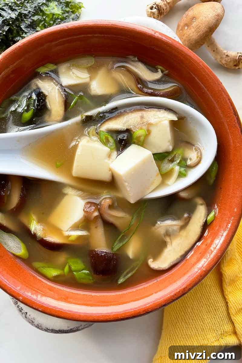 Vertical image of a spoon over a bowl of broth with mushrooms, tofu, and scallions next to a yellow towel.