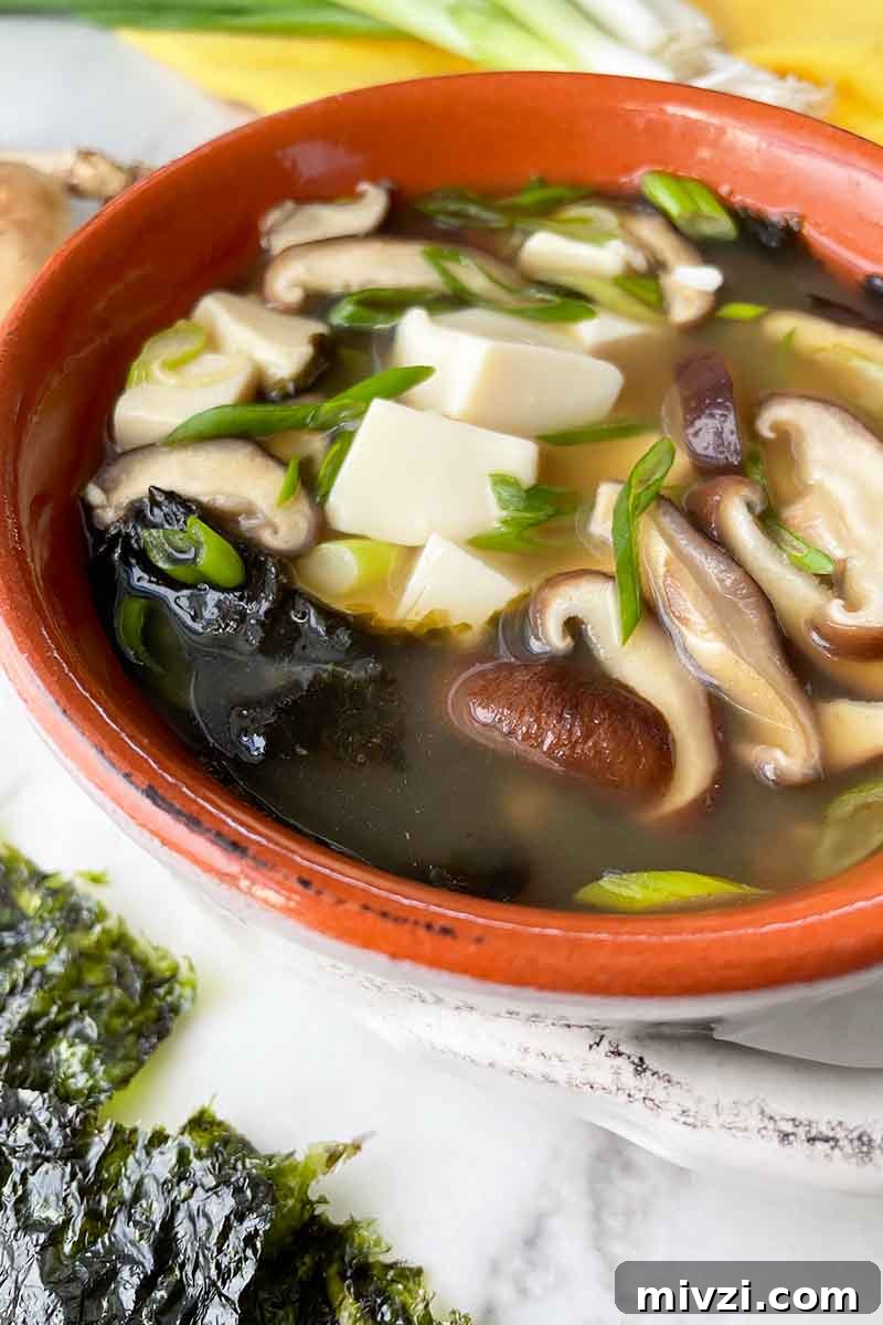 Vertical close-up image of a bowl full of broth with tofu chunks, sliced shiitake, and seaweed pieces.