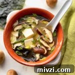 Horizontal image of a red bowl filled with broth with chunks of tofu, sliced mushrooms, and seaweed with a spoon inserted into it, next to a green towel