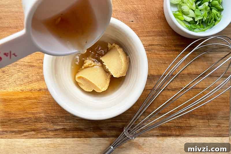Horizontal image of pouring water over a thick paste in a white bowl next to a whisk and a bowl of scallions.
